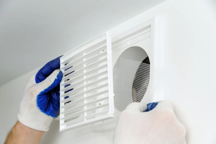 Plombier professionnel en tenue de travail effectuant une installation de gaine VMC au plafond d'une maison, à côté d'un client qui observe et communique avec lui. La scène montre le plombier en action dans un intérieur résidentiel, illustrant l'expertise technique et la relation directe avec le client dans un environnement réel d'installation.