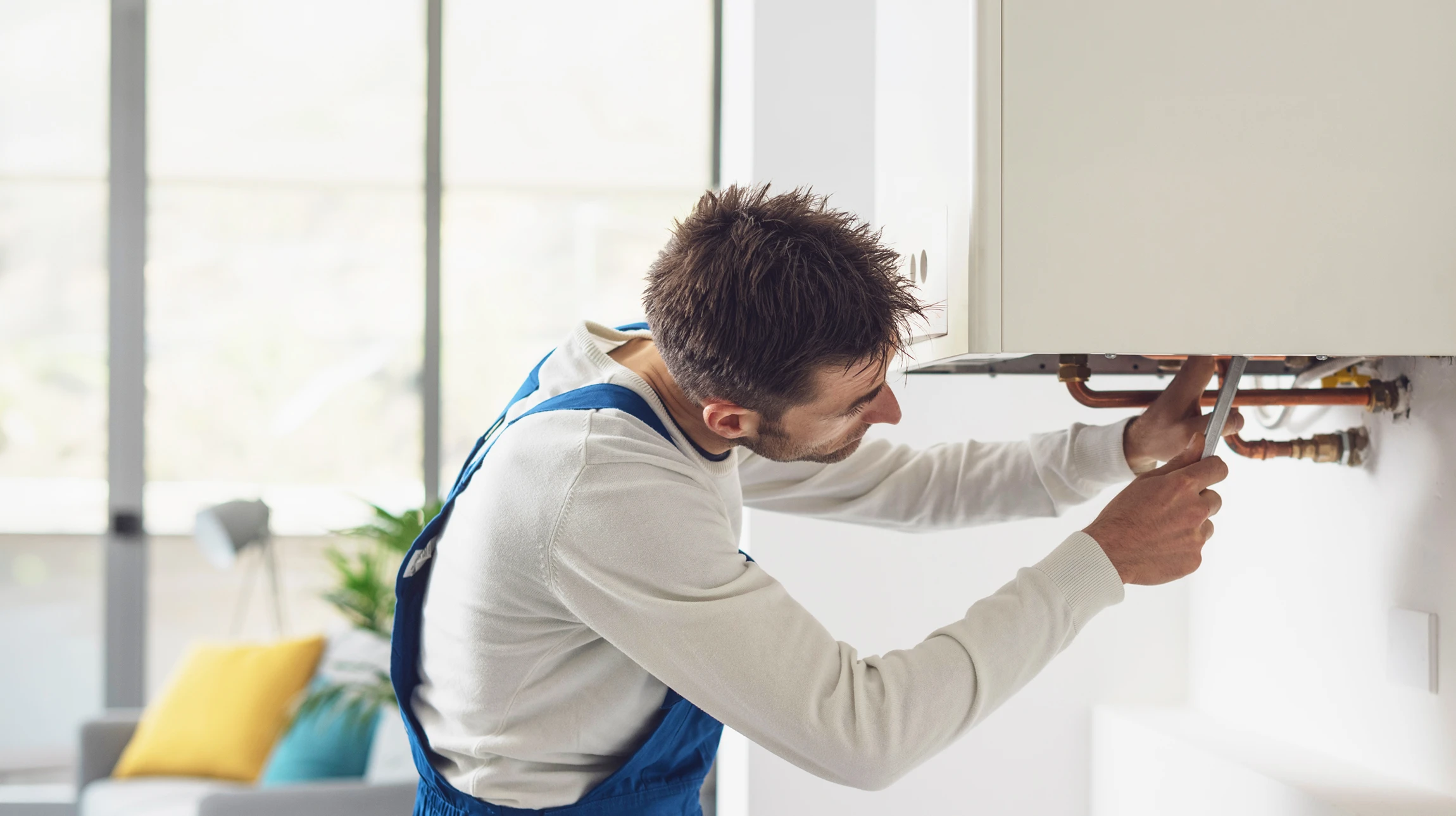 Photographie r&eacute;aliste d'un artisan plombier professionnel dans un int&eacute;rieur r&eacute;sidentiel, effectuant un diagnostic ou une intervention sur une tuyauterie/radiateur. L'artisan porte une tenue de travail appropri&eacute;e (v&ecirc;tements de m&eacute;tier, outils &agrave; la ceinture ou &agrave; proximit&eacute;). La sc&egrave;ne doit montrer une action concr&egrave;te li&eacute;e au d&eacute;pannage ou la r&eacute;novation sanitaire/chauffage, dans un environnement d'habitation authentique, photographi&eacute;e sous un &eacute;clairage naturel professionnel.