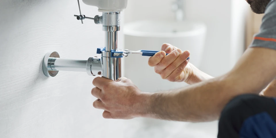 Photographie d'un artisan plombier en tenue professionnelle travaillant &agrave; l'int&eacute;rieur d'une salle de bain en r&eacute;novation, montrant &agrave; la fois des outils de d&eacute;pannage urgent (d&eacute;boucheur, cl&eacute; anglaise) et les phases finales d'installation sanitaire (raccordement de radiateur, finitions de robinetterie). L'image doit transmettre la polyvalence et la ma&icirc;trise technique de l'artisan capable de g&eacute;rer l'urgence et la r&eacute;novation compl&egrave;te en un m&ecirc;me lieu.